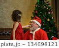 Young child girl in red dress feeding cookie to santa claus in living room near decorated Christmas tree with ornaments and twinkling lights at home 134698121