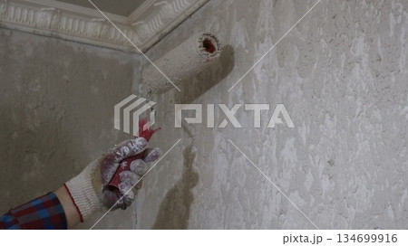 The left hand in a glove holds a roller and applies mortar, putty, primer or paint to the wall under the ceiling, repair work in the room, preparing interior walls for renovation 134699916