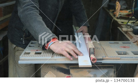 A carpenter saws a long narrow board on a circular saw in an open-plan workshop, working with wood on a machine tool, processing and preparing building material in a carpentry shop A carpenter saws a long narrow board on a circular saw in an open-plan workshop, working with wood on a machine tool, processing and preparing building material in a carpentry shop 134700138