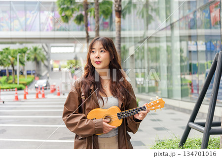 Young woman happy person travel tourist enjoys holiday music, playing ukulele outdoors on vibrant urban city street, embracing summer lifestyle with joyful expression, walking among modern buildings 134701016