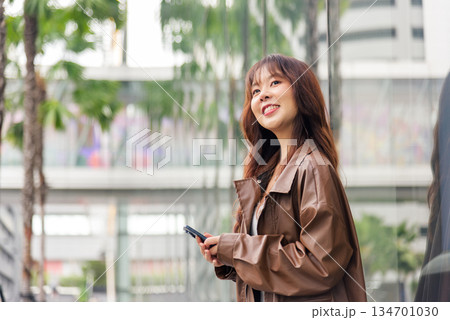 Asian woman enjoys shopping lifestyle, fashion bags complement urban mall experience, people explore vibrant city scene, smiling, holding smartphone, wearing stylish brown jacket, modern casual look 134701030