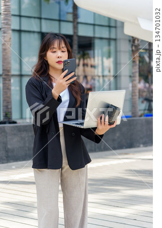 Young Asian business woman, busy professional, on city street works, wearing suit, managing digital tasks using smartphone, holding laptop, standing outdoor in urban environment communication Young Asian business woman, busy professional, on city street works, wearing suit, managing digital tasks using smartphone, holding laptop, standing outdoor in urban environment communication 134701032