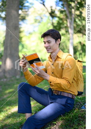 A man from an office sits on the grass in a green park reading a book with a relaxed mood 134703304