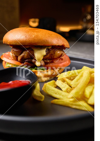 Gourmet Cheeseburger with Juicy Chicken, Fresh Vegetables, Crispy Fries, and Ketchup on Black Plate, Delicious Fast Food Meal Concept 134703469