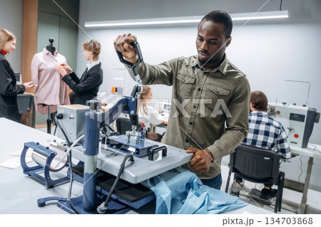 Black man is standing by the table. People are working by the sewing machines indoors Black man is standing by the table. People are working by the sewing machines indoors 134703868