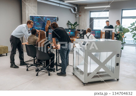 Big screen with code on the wall. People are in the office by the 3d printer 134704632