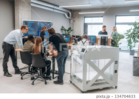 Big screen with code on the wall. People are in the office by the 3d printer 134704633