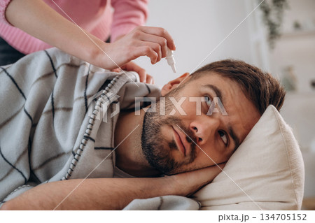 Funny facial expression. A man lies on a bed. A woman puts ear drops in his ear Funny facial expression. A man lies on a bed. A woman puts ear drops in his ear 134705152