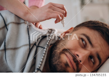 Funny facial expression. A man lies on a bed. A woman puts ear drops in his ear Funny facial expression. A man lies on a bed. A woman puts ear drops in his ear 134705155