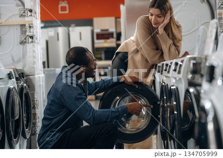 Sitting and opening. Man and woman are choosing washing machine in the store Sitting and opening. Man and woman are choosing washing machine in the store 134705999