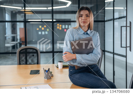 Posing, sitting on the table, holding laptop. Woman is in the modern office, formal wear Posing, sitting on the table, holding laptop. Woman is in the modern office, formal wear 134706080