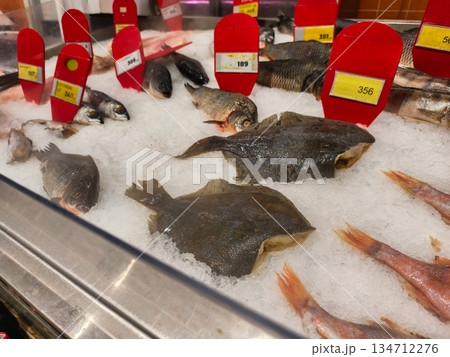 Fish on display. A variety of fresh seafood, including whole flounder, perch, and red mullet, is displayed on crushed ice with red price tags at a supermarket or fish market. 134712276