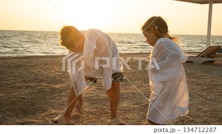 A brother and sister explore the coastline during golden hour, barefoot on the sand. The warm sunset light and calm sea create a peaceful, carefree picture of a summer evening as the children play. 134712287