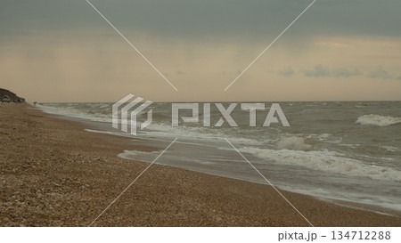 Under a gloomy, overcast sky, stormy sea waves crash against a pebble beach, creating a dramatic coastal landscape that embodies the power and unpredictability of nature. A storm at sea. 134712288