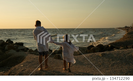 Two children, a boy and a girl, holding hands and walking together on a rocky beach towards the ocean during a warm sunset, symbolizing family connection, childhood, and summer freedom. 134712290