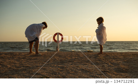 One child watches another child playing with a stick on the beach. They stand at sunset, separated by a life preserver, symbolizing safety and distance. Children playing on the beach at sunset. One child watches another child playing with a stick on the beach. They stand at sunset, separated by a life preserver, symbolizing safety and distance. Children playing on the beach at sunset. 134712291