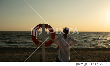 A boy stands on a pebble beach looking out at the calm ocean during a golden sunset. A teenager stands near a lifebuoy on the seashore at sunset. A child at sunset against the backdrop of a calm sea. A boy stands on a pebble beach looking out at the calm ocean during a golden sunset. A teenager stands near a lifebuoy on the seashore at sunset. A child at sunset against the backdrop of a calm sea. 134712292