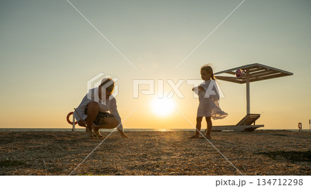 Children explore the sandy shore during golden hour. Their silhouettes stand out against the warm sunset over the calm ocean, evoking memories of carefree and peaceful family moments. 134712298