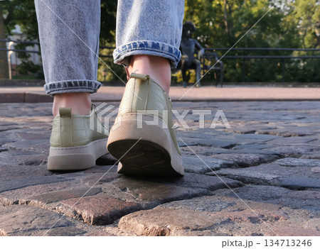 Close-up of someones feet in sneakers stepping on a cobblestone path, with a blurred statue in the background. Close-up of someones feet in sneakers stepping on a cobblestone path, with a blurred statue in the background. 134713246