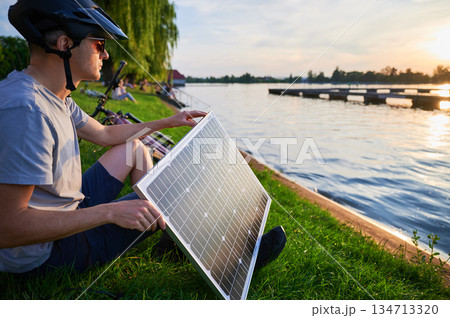 Man cyclist using solar panel for charging electric mountain bike outdoors. Guy in helmet and sunglasses sits by lakeside, enjoying tranquil scene as sun sets over water. Man cyclist using solar panel for charging electric mountain bike outdoors. Guy in helmet and sunglasses sits by lakeside, enjoying tranquil scene as sun sets over water. 134713320