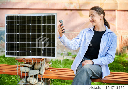 Happy woman using smartphone connected to photovoltaic solar panel. Integration of sustainable renewable energy into everyday life, demonstrating practical use of solar power for charging devices. 134713322