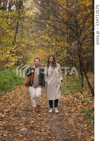 Two women walk together along a forest path covered with autumn leaves, tall trees around, natural daylight, relaxed and friendly atmosphere 134713670