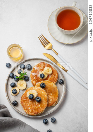 Pancakes with blueberry and banana on a plate on a light background with cup of tea Pancakes with blueberry and banana on a plate on a light background with cup of tea 134713832