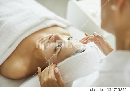 Female beautician applying moisturizing sheet mask on young woman face in beauty spa salon. 134713852