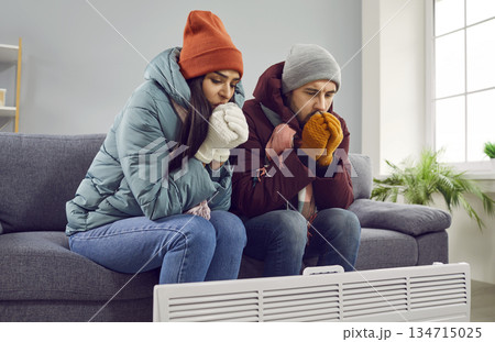 Young attractive couple are sitting on the sofa at home in warm clothes and warming their hands with heater in the living room, freezing from the cold. Heating problems, room is cold, people freezing. Young attractive couple are sitting on the sofa at home in warm clothes and warming their hands with heater in the living room, freezing from the cold. Heating problems, room is cold, people freezing. 134715025