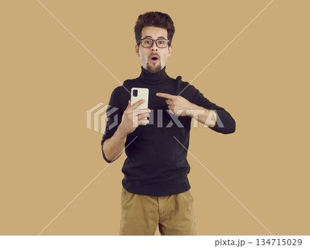 Amazed young brunet man in black turtleneck is pointing at smartphone on beige background. Amazed young brunet man in black turtleneck is pointing at smartphone on beige background. 134715029