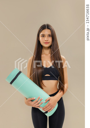 Young brunette woman in black sportswear holds a turquoise yoga mat, posing against a beige background, fitness and healthy lifestyle concept. 134716638