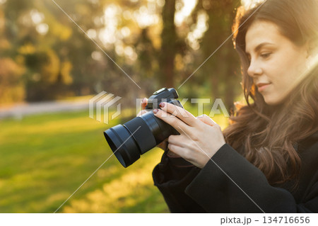 Close-up of a young woman holding a digital camera while photographing outdoors in warm sunlight. Soft golden-hour light, creative lifestyle photography moment. Close-up of a young woman holding a digital camera while photographing outdoors in warm sunlight. Soft golden-hour light, creative lifestyle photography moment. 134716656