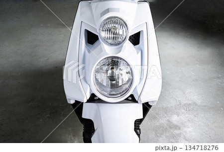 Front view of white motorcycle or scooter with round headlights and modern design parked indoors showing symmetry transport concept and urban mobility Front view of white motorcycle or scooter with round headlights and modern design parked indoors showing symmetry transport concept and urban mobility 134718276