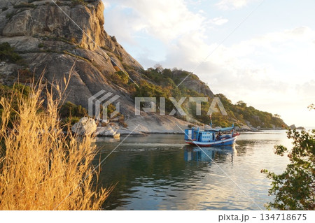 A colorful wooden boat sets out to sea at dawn to fish. It's morning, dawn, and hot. In the background is a mountain with green trees and grass and a blue sky.A fishing boat going out to sea to fish. 134718675