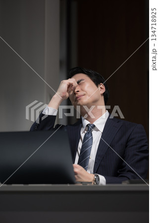 暗いオフィスで仕事が終わらず残業でパソコンの前で眼精疲労や頭痛のビジネスマン 134718925