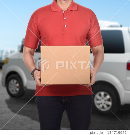 Delivery person in a red polo shirt stands holding a brown cardboard box, ready to deliver the package. A white van is visible in the background, creating a scene of delivery services 134719921