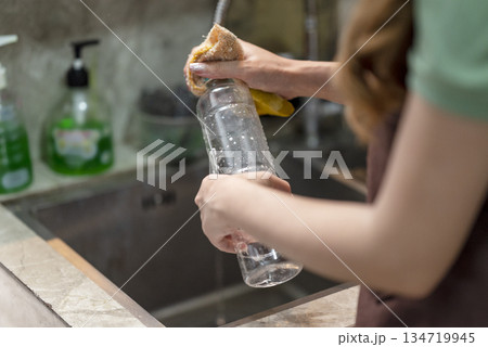 Person hands washing a clear plastic bottle with a yellow sponge under a stream of water in a kitchen sink, promoting cleanliness and hygiene Person hands washing a clear plastic bottle with a yellow sponge under a stream of water in a kitchen sink, promoting cleanliness and hygiene 134719945