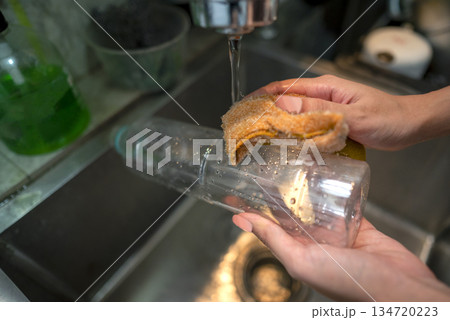 Person hands washing a clear plastic bottle with a yellow sponge under a stream of water in a kitchen sink, promoting cleanliness and hygiene Person hands washing a clear plastic bottle with a yellow sponge under a stream of water in a kitchen sink, promoting cleanliness and hygiene 134720223