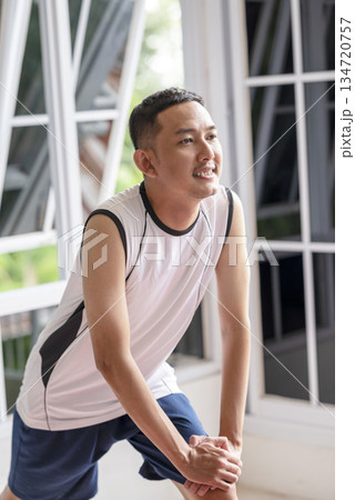 Indonesian southeast asian man in activewear stretching his leg indoors. Morning routine exercise, showcasing a healthy lifestyle Indonesian southeast asian man in activewear stretching his leg indoors. Morning routine exercise, showcasing a healthy lifestyle 134720757
