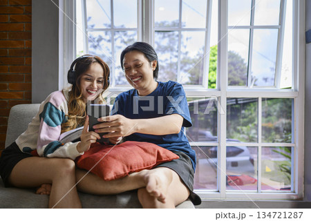 Indonesian southeast asian couple is sitting together on a couch, while looking at something on a cellphone. They are both smiling and enjoying, creating a warm and relatable atmosphere 134721287