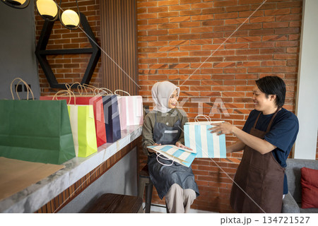 Indonesian southeast asian muslim woman and a man in an apron holding a striped shopping bag inside a modern store with colorful bags displayed on the counter Indonesian southeast asian muslim woman and a man in an apron holding a striped shopping bag inside a modern store with colorful bags displayed on the counter 134721527