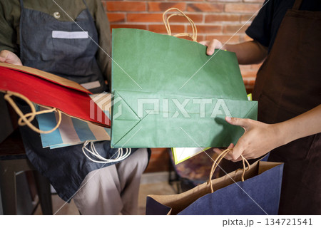 Indonesian southeast asian muslim woman and a man in an apron holding a striped shopping bag inside a modern store with colorful bags displayed on the counter Indonesian southeast asian muslim woman and a man in an apron holding a striped shopping bag inside a modern store with colorful bags displayed on the counter 134721541