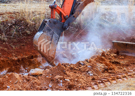 Excavator operates on dirt path, removing soil in ground during digging trench work 134722316