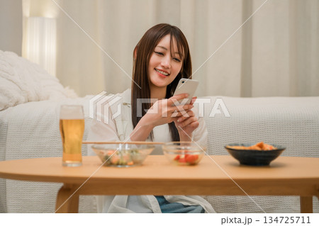 家の寝室で晩酌しながらスマホを見る若いアジア人女性（ビール,家飲み,飲酒習慣） 134725711