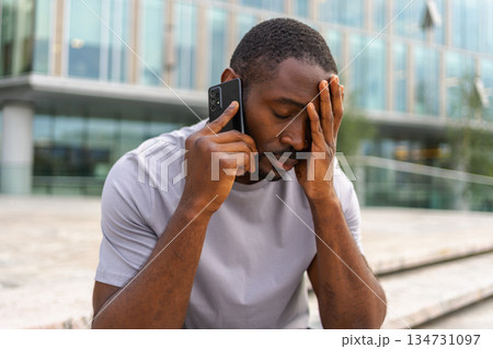 Sad tired lonely disappointed African man alone talking on smartphone on city street outdoor. Unhappy person making answering call by cellphone experiencing grief getting bad news. Stressed man 134731097