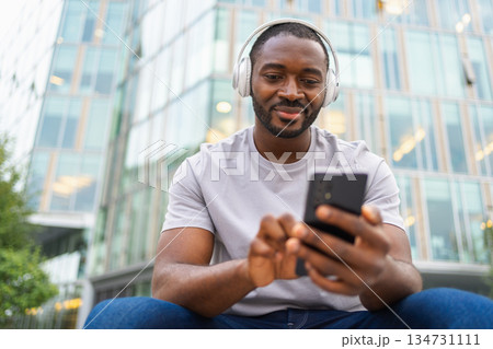 Music song concept. Good start day. Happy African American man listening modern hit on street in city. Person guy wearing headphones enjoy listening favorite music on phone. People lifestyle joy Music song concept. Good start day. Happy African American man listening modern hit on street in city. Person guy wearing headphones enjoy listening favorite music on phone. People lifestyle joy 134731111