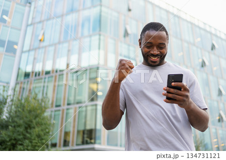 Happy African American man euphoric winner with smartphone on street in city. Person guy looking at cell phone reading great news getting good result winning online bid feeling amazed. Winning gesture Happy African American man euphoric winner with smartphone on street in city. Person guy looking at cell phone reading great news getting good result winning online bid feeling amazed. Winning gesture 134731121
