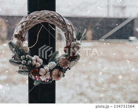 Cozy winter holiday scene. Handmade Christmas wreath hanging on a window pane with snow falling outdoors, festive mood, domestic life, seasonal warmth Cozy winter holiday scene. Handmade Christmas wreath hanging on a window pane with snow falling outdoors, festive mood, domestic life, seasonal warmth 134731564