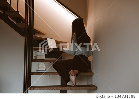 Young woman working on laptop at home office sitting on wooden stairs, cozy minimalist interior, remote work and creative freelance lifestyle concept, calm productivity and modern slow living vibe. 134731589