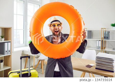 Joyful happy man holding yellow suitcase and swim ring while standing in office Joyful happy man holding yellow suitcase and swim ring while standing in office 134735014